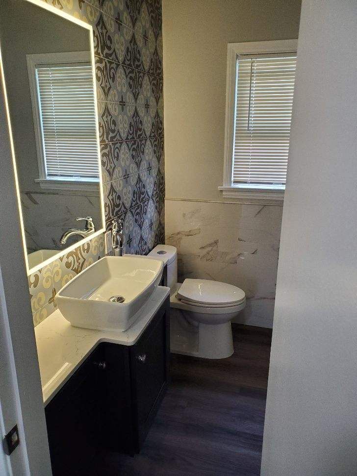 Bathroom with vanity, toilet, patterned wallpaper, and wood-look flooring. Window with blinds.