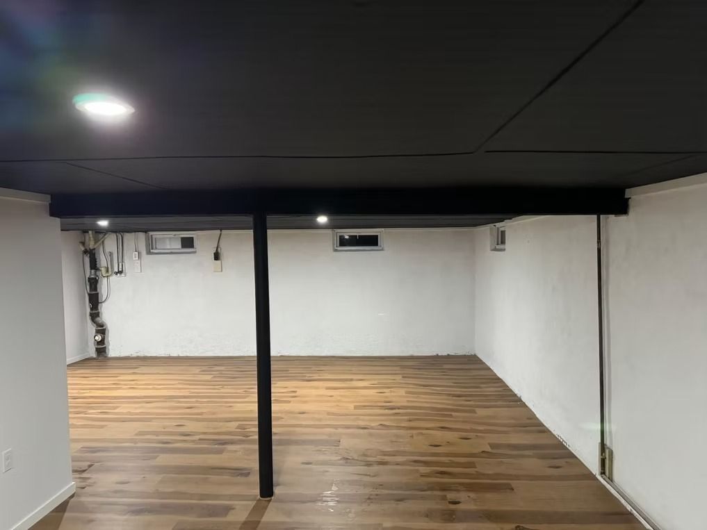 Empty basement room with white walls, wood floor, black ceiling, and a central support post