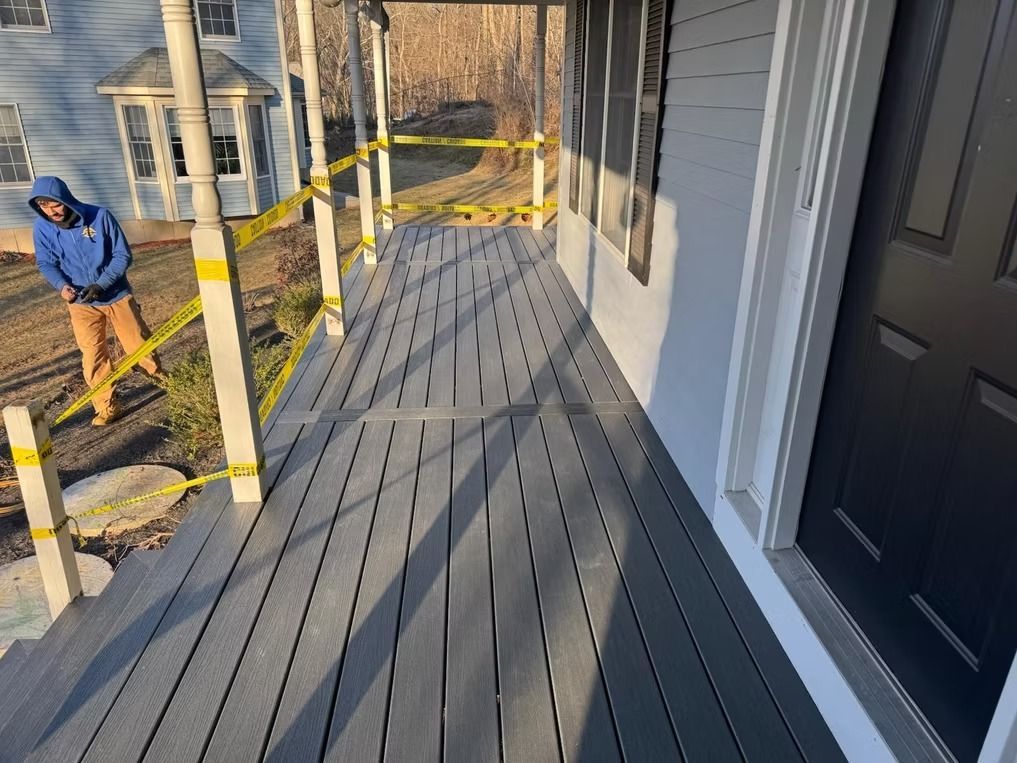 Gray deck under construction beside a house, with yellow caution tape and a worker in a blue hoodie nearby