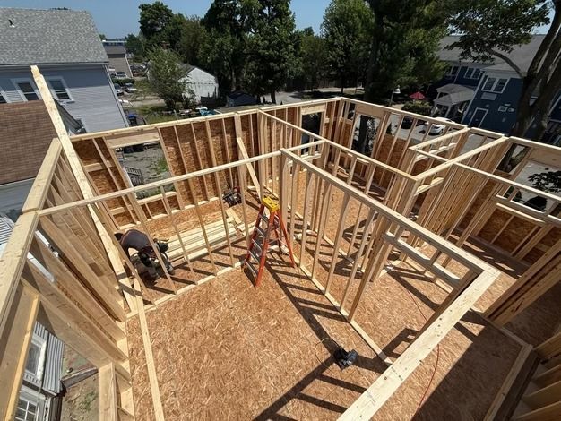 Wood-framed house under construction, with two workers on an OSB floor and a ladder in the center.