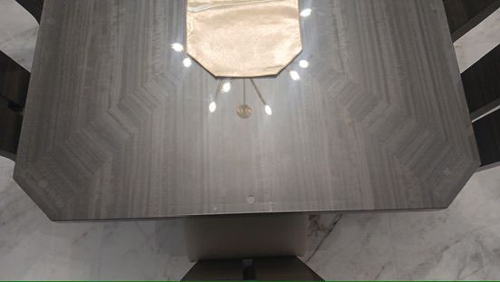A top-down view of a rectangular, grey-toned wooden dining table with a neutral-colored runner, reflecting overhead lights.