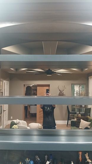 A person stands in a room with a vaulted ceiling, visible through the reflection of three horizontal glass shelves.