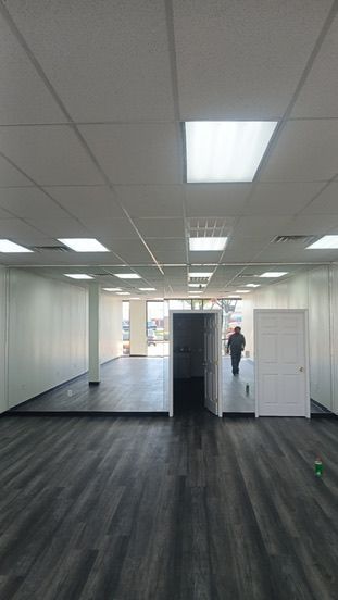 An empty, modern commercial space with dark wood-look flooring, white walls, drop ceiling lights, and a white door.