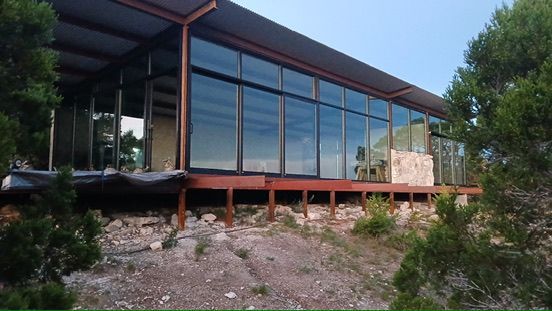 Modern house with floor-to-ceiling windows and a wooden deck, set on a rocky, shrub-covered slope under a clear sky.
