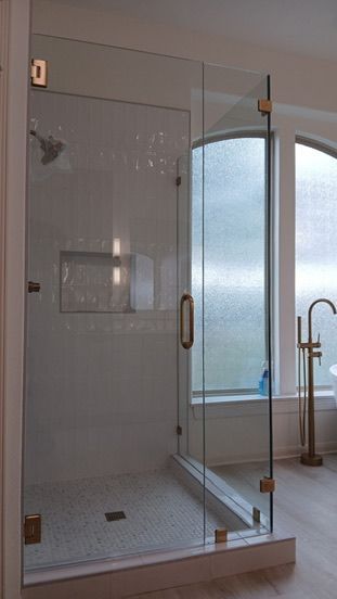 A glass-enclosed shower with gold hardware, a white tile floor, and a gold freestanding faucet beside a window.