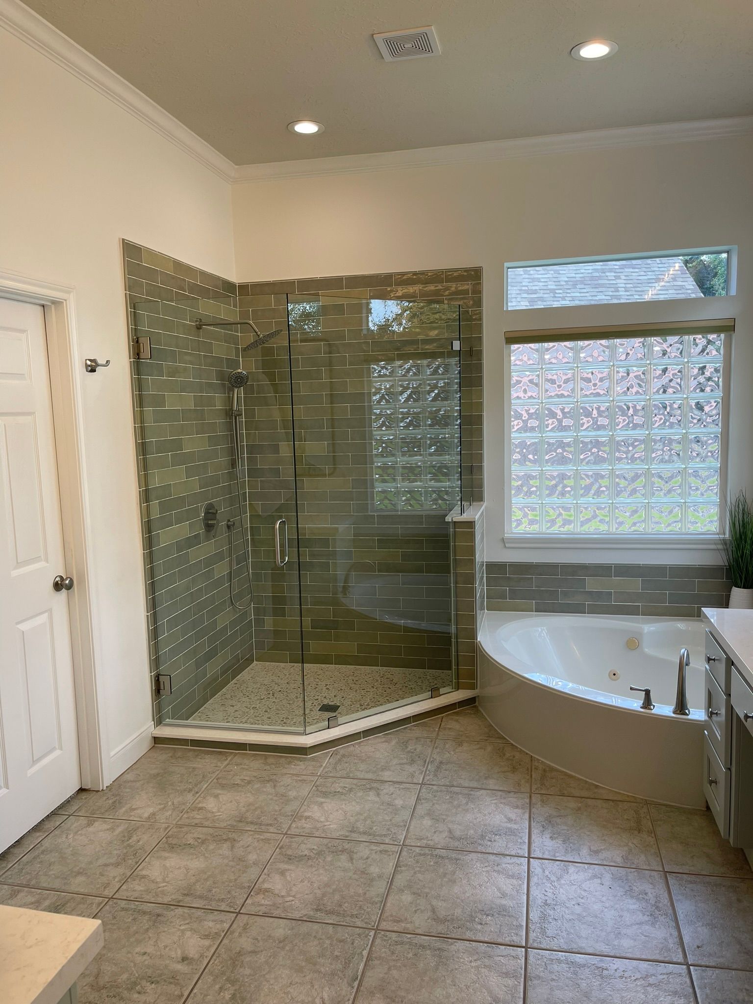 A spacious bathroom featuring a tiled walk-in glass shower, a jetted corner tub, and a window with glass privacy blocks.