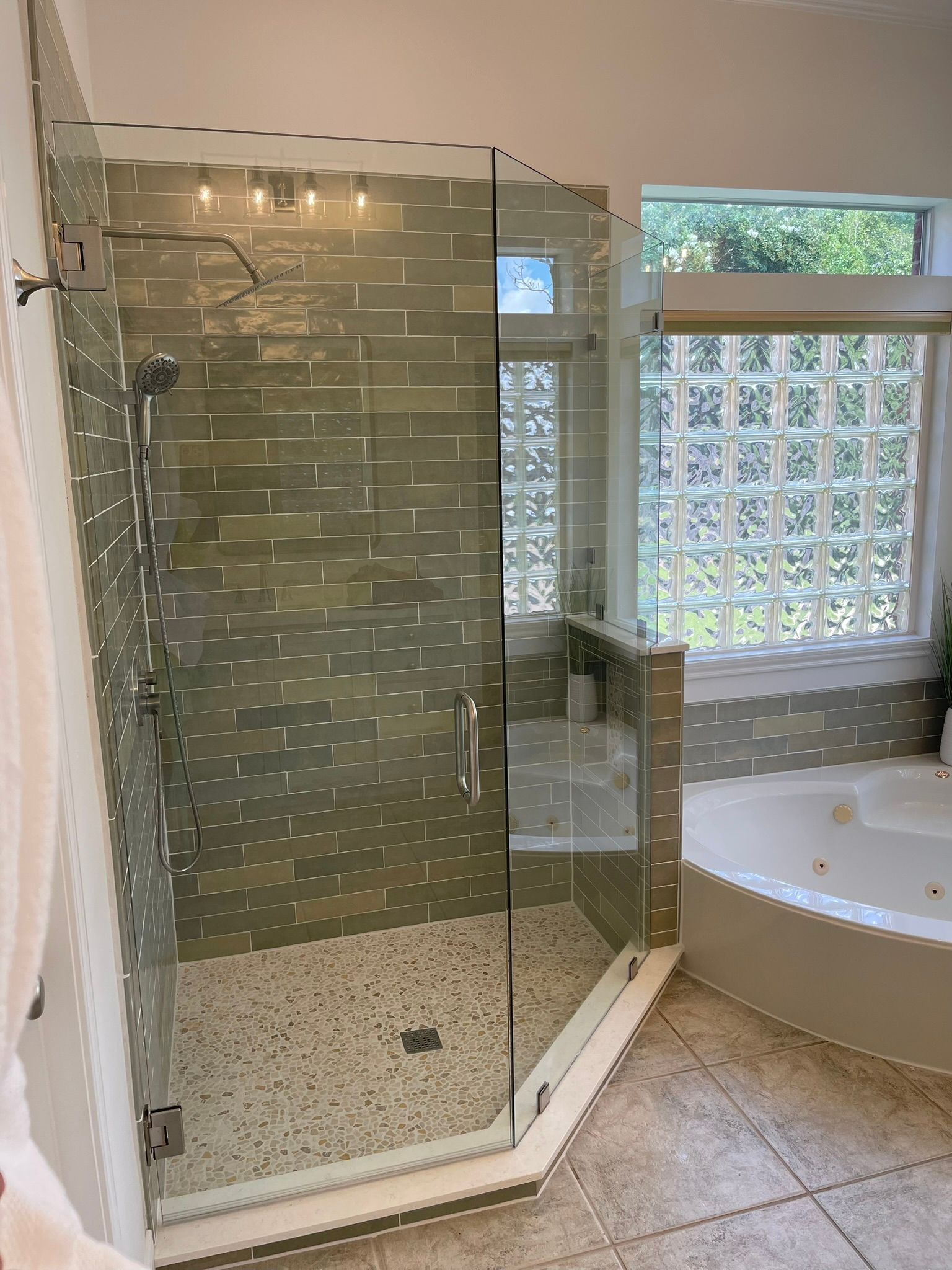 A glass-enclosed shower with olive stone tiles and pebble flooring next to a jetted bathtub in a bright bathroom.