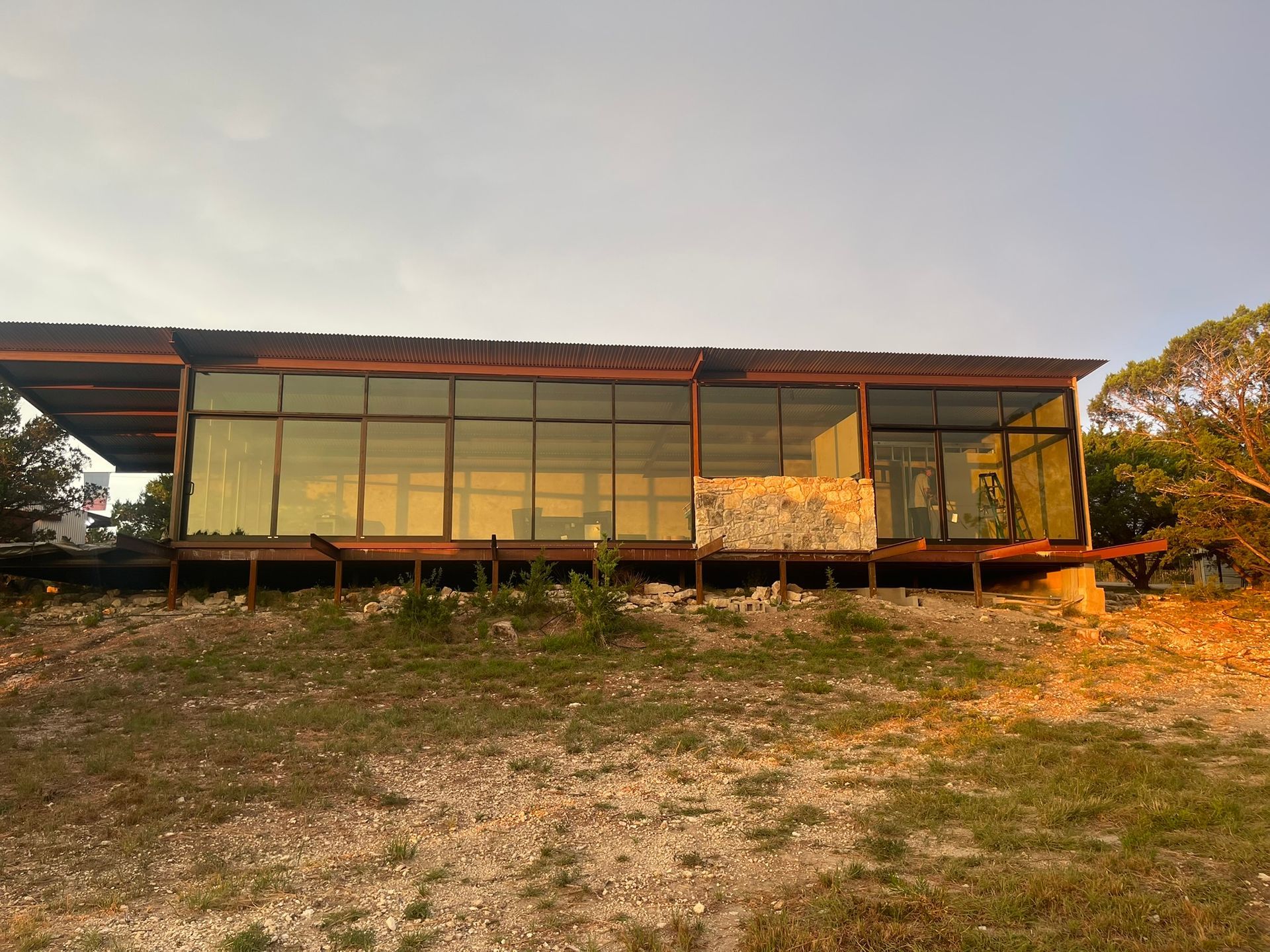 A modern, long, rectangular building with floor-to-ceiling glass walls stands on a rocky hill under a soft sunset sky.