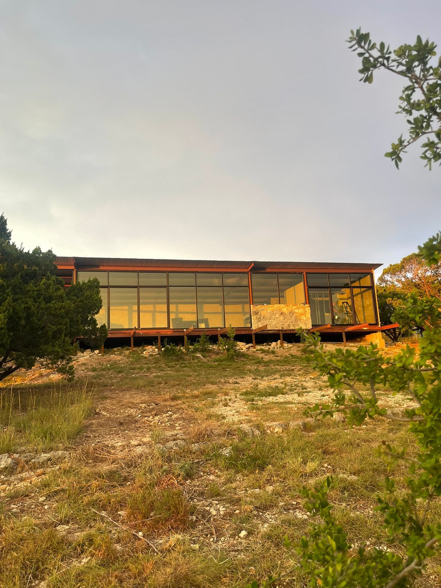 A modern, flat-roofed house with floor-to-ceiling windows sits atop a rocky, grassy hill under a bright, cloudy sky.