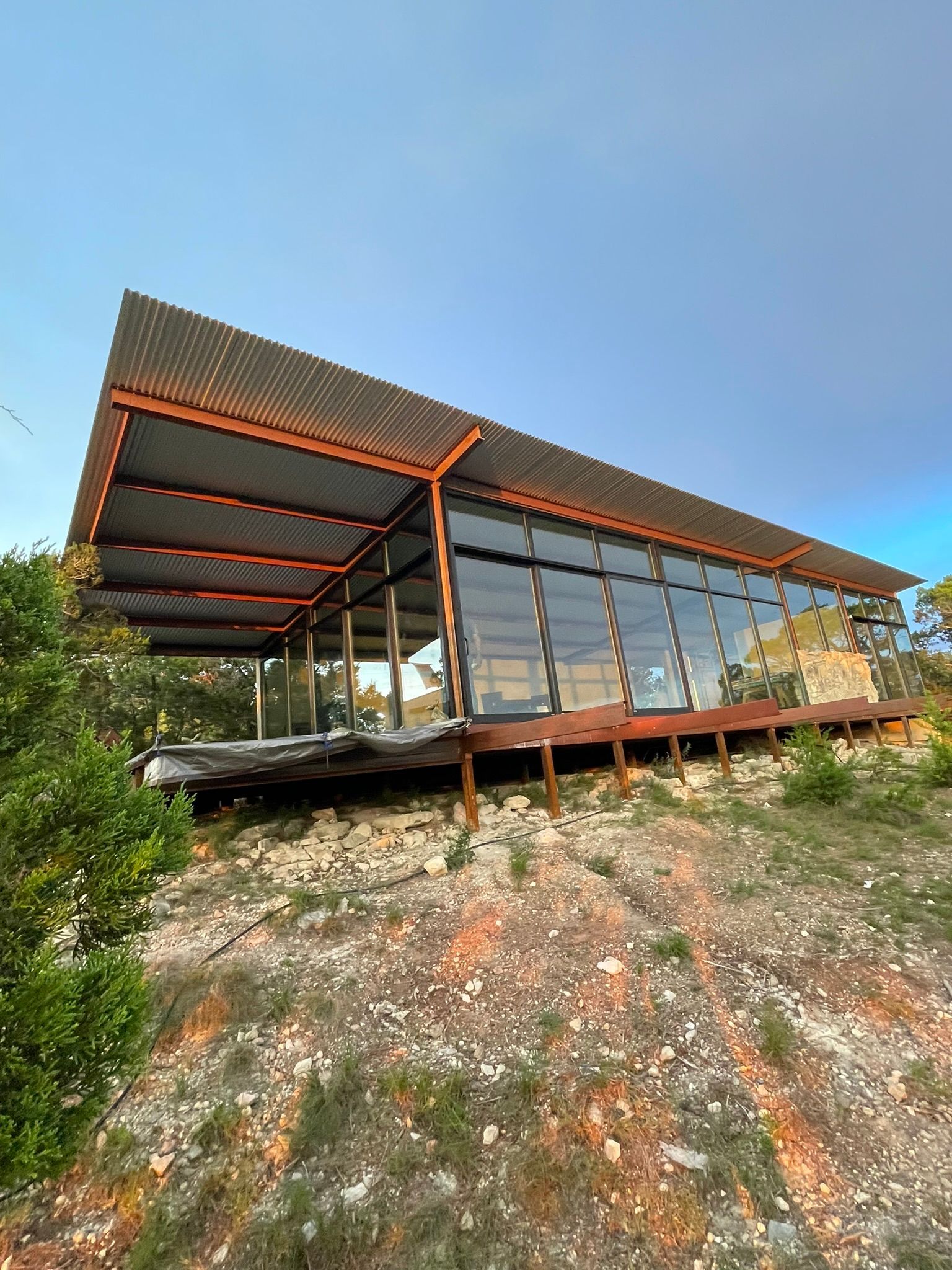 A modern building with a sloped metal roof and large glass walls stands on a rocky hillside under a clear blue sky.