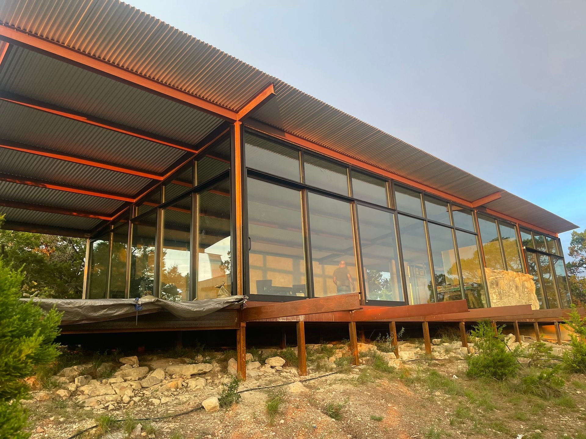 A modern, rectangular cabin with large glass walls and a corrugated metal roof, raised on stilts above rocky ground.