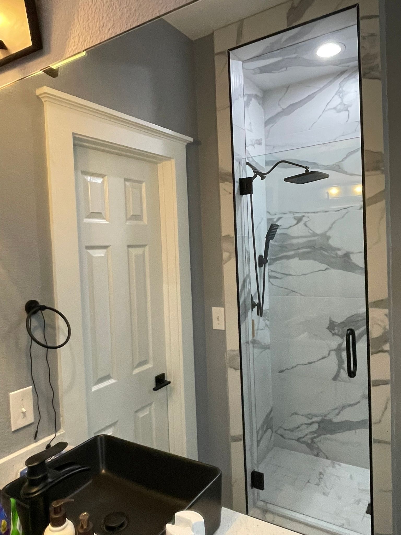 A bathroom vanity with a black sink and towel ring next to a marble-tiled walk-in shower with a glass door.