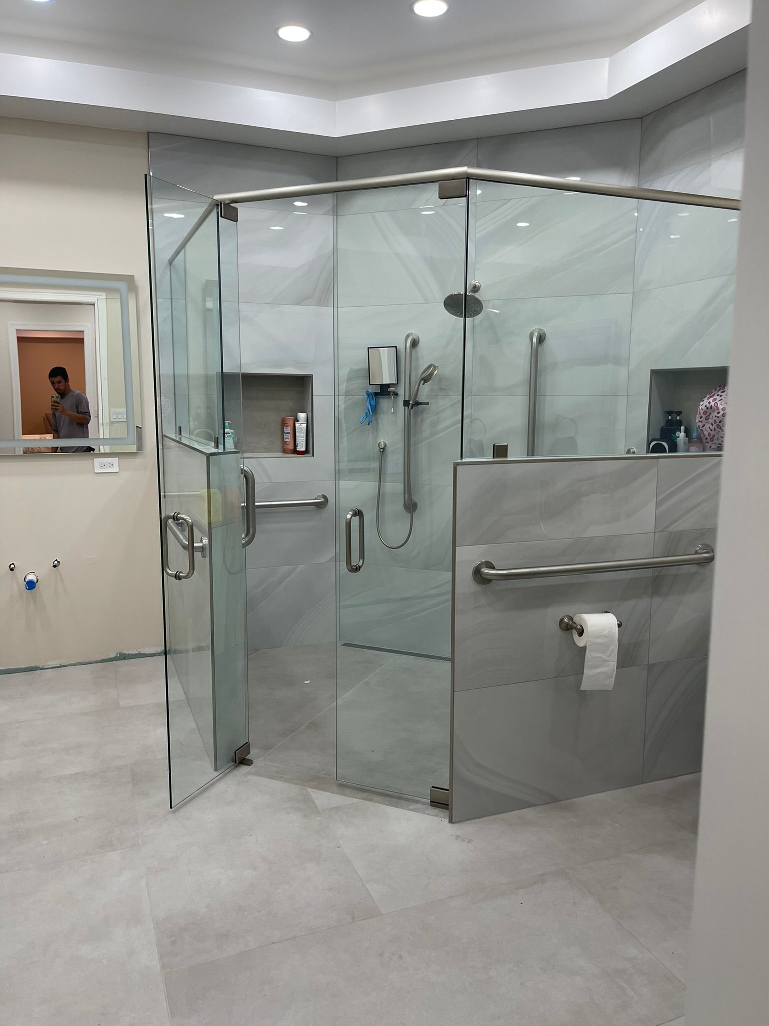 A modern, light gray tiled walk-in shower with glass doors, silver grab bars, and a toilet paper holder mounted on the wall.