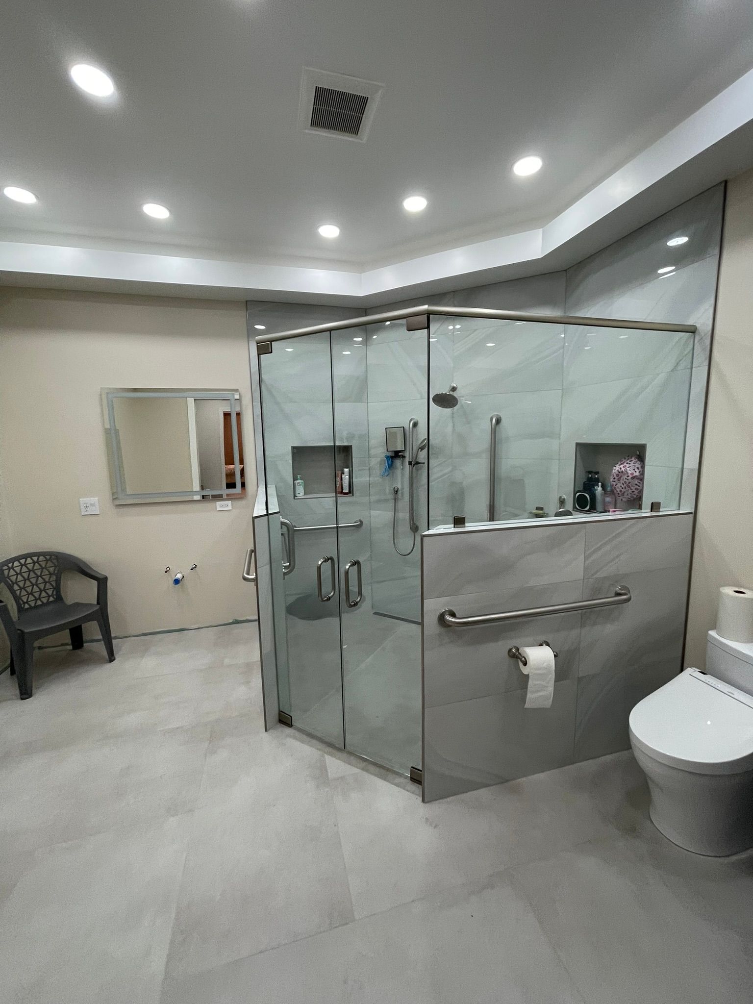 A modern bathroom with a glass-enclosed shower, a toilet with a grab bar, large neutral floor tiles, and a gray chair.