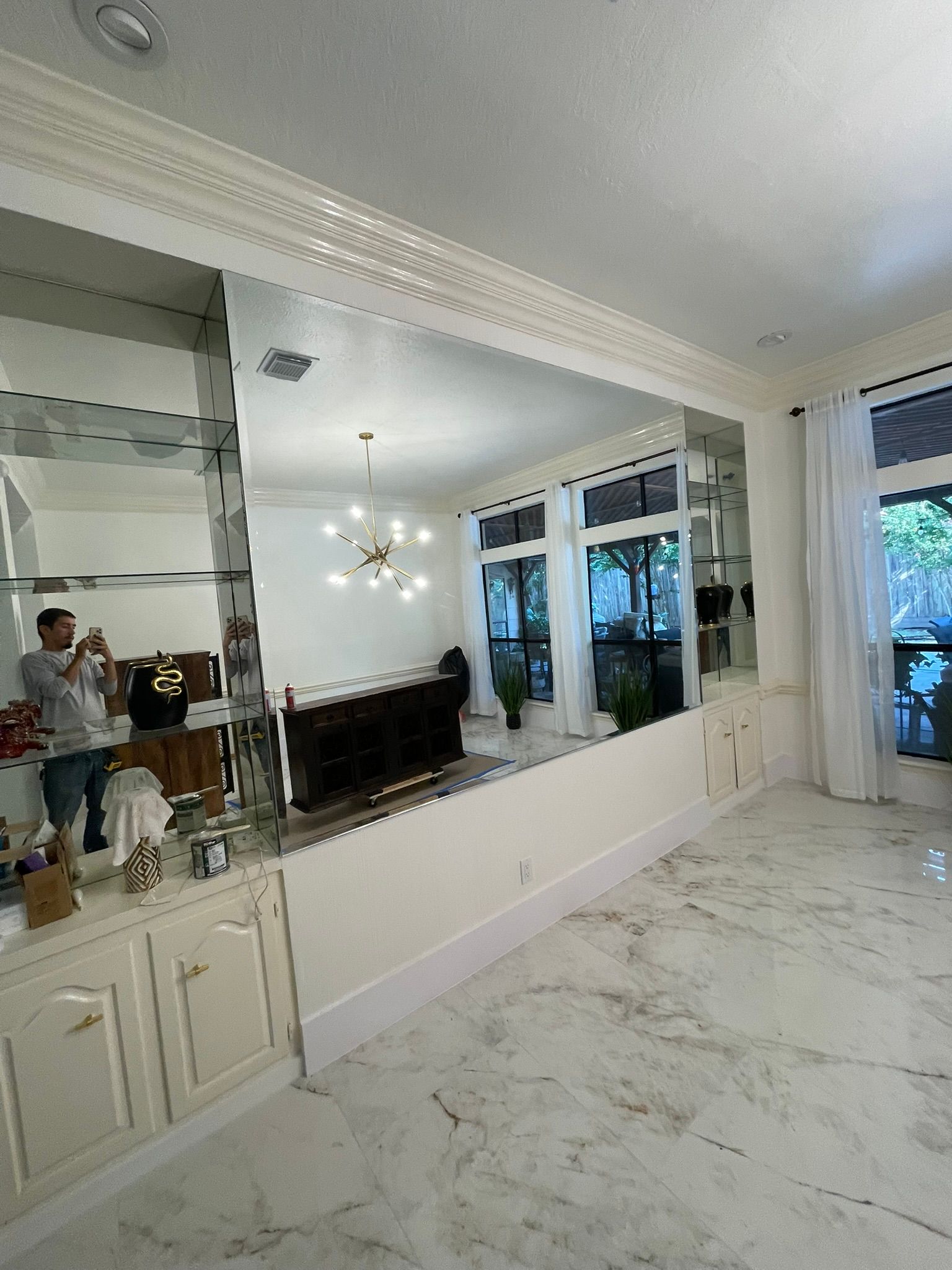 A person taking a photo in a large, wall-to-wall mirror within a room with marble floors and white cabinetry.