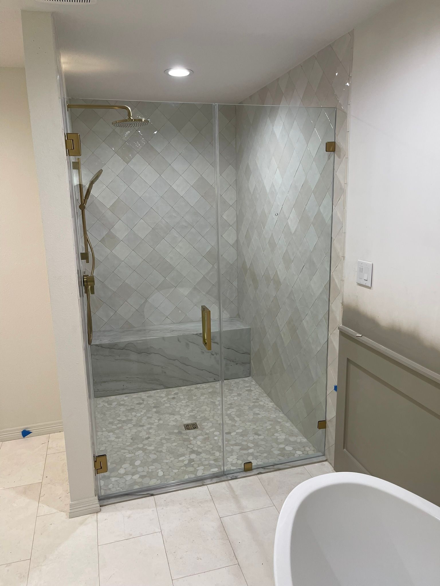 A glass-enclosed shower with patterned white tile walls, a stone bench, pebble floor, and gold hardware in a bathroom.