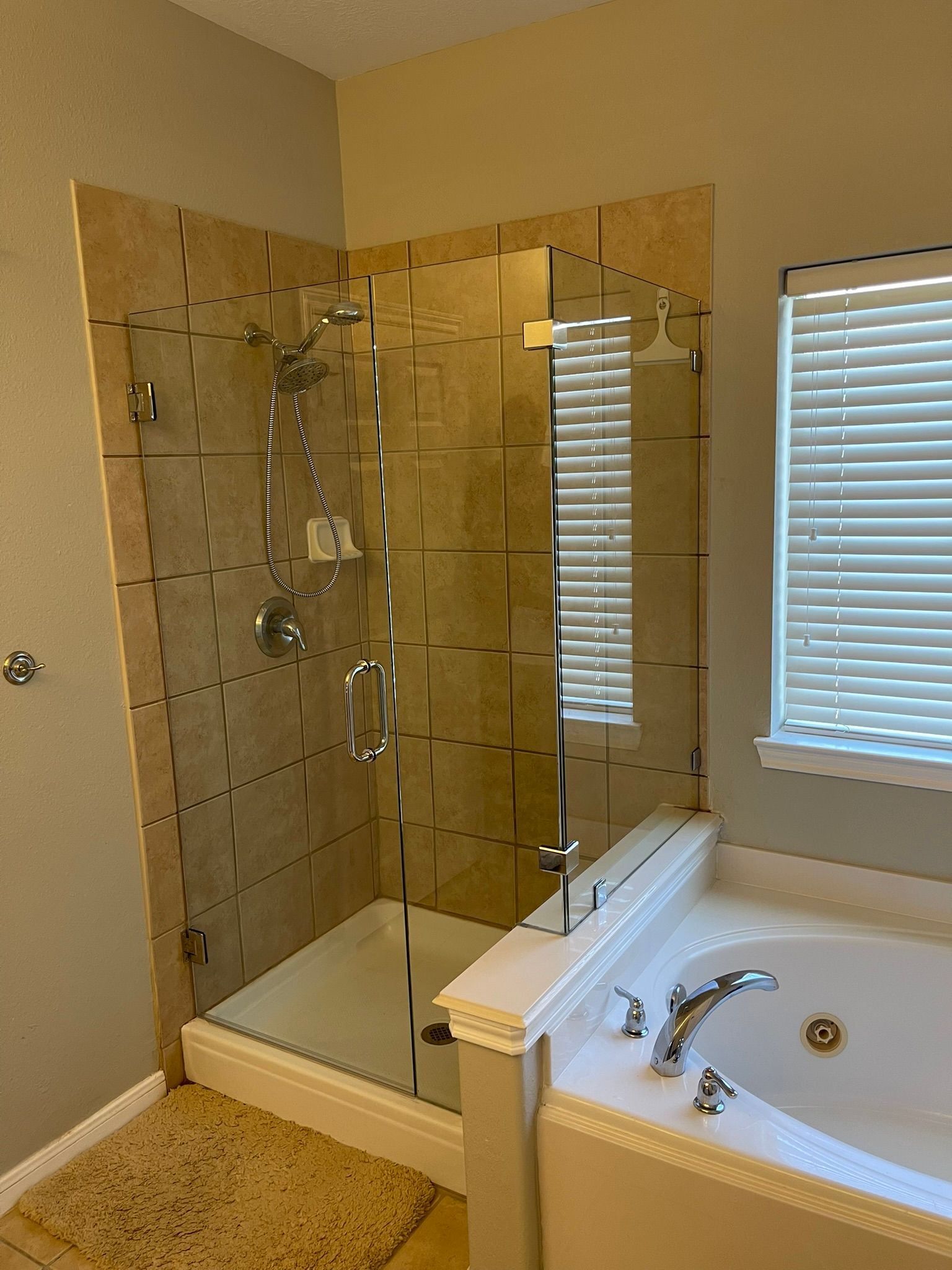 A glass-enclosed shower with beige tile walls next to a white bathtub in a bathroom with window blinds.
