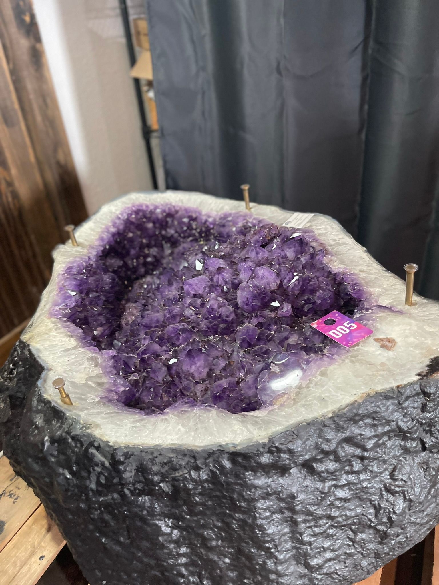 A large, deep purple amethyst geode with a dark, textured rock exterior, displayed on a wooden stand indoors.