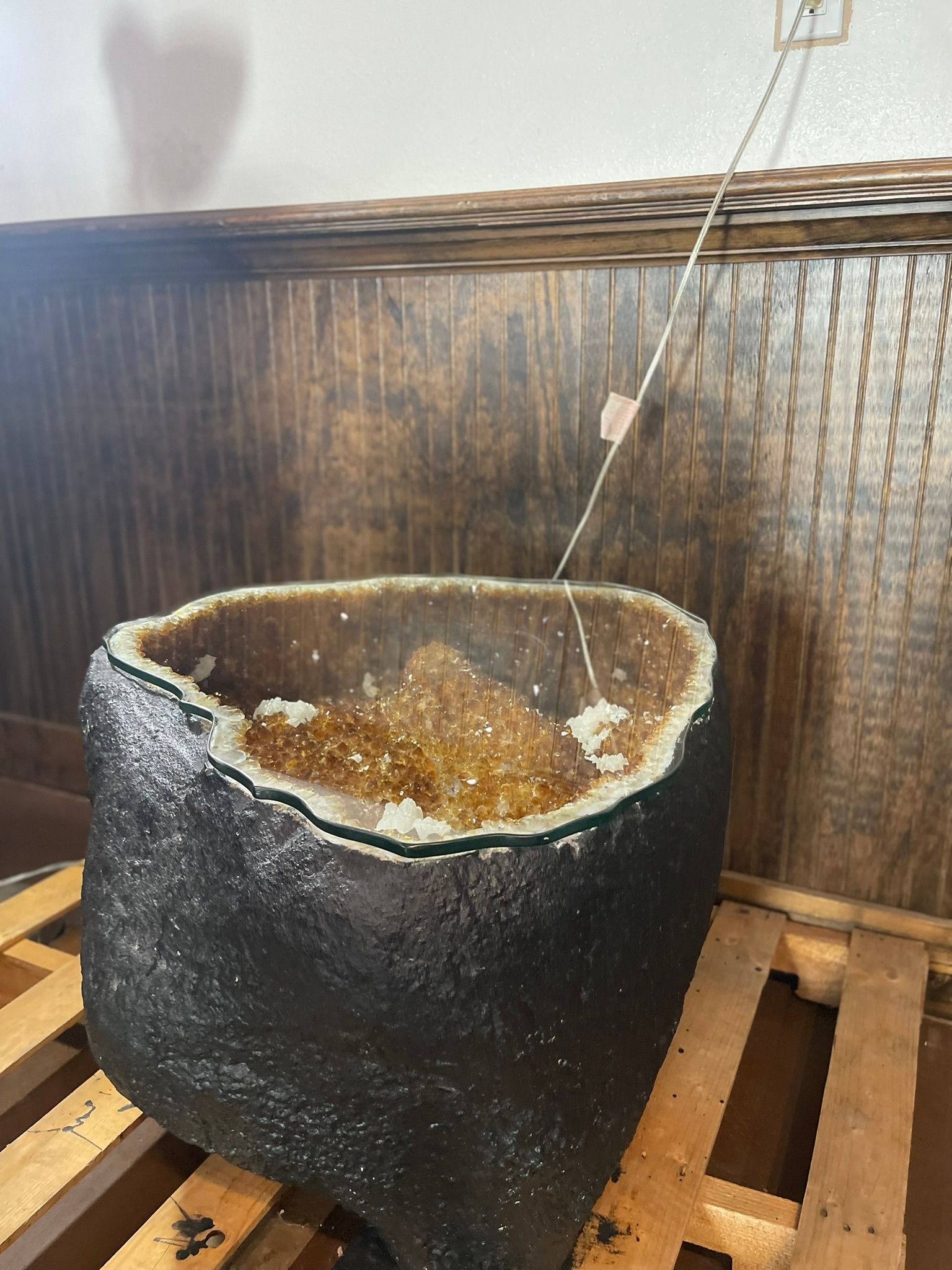 A large, dark, rough-textured geode with amber-colored interior crystals sitting on a wooden pallet against a wood wall.