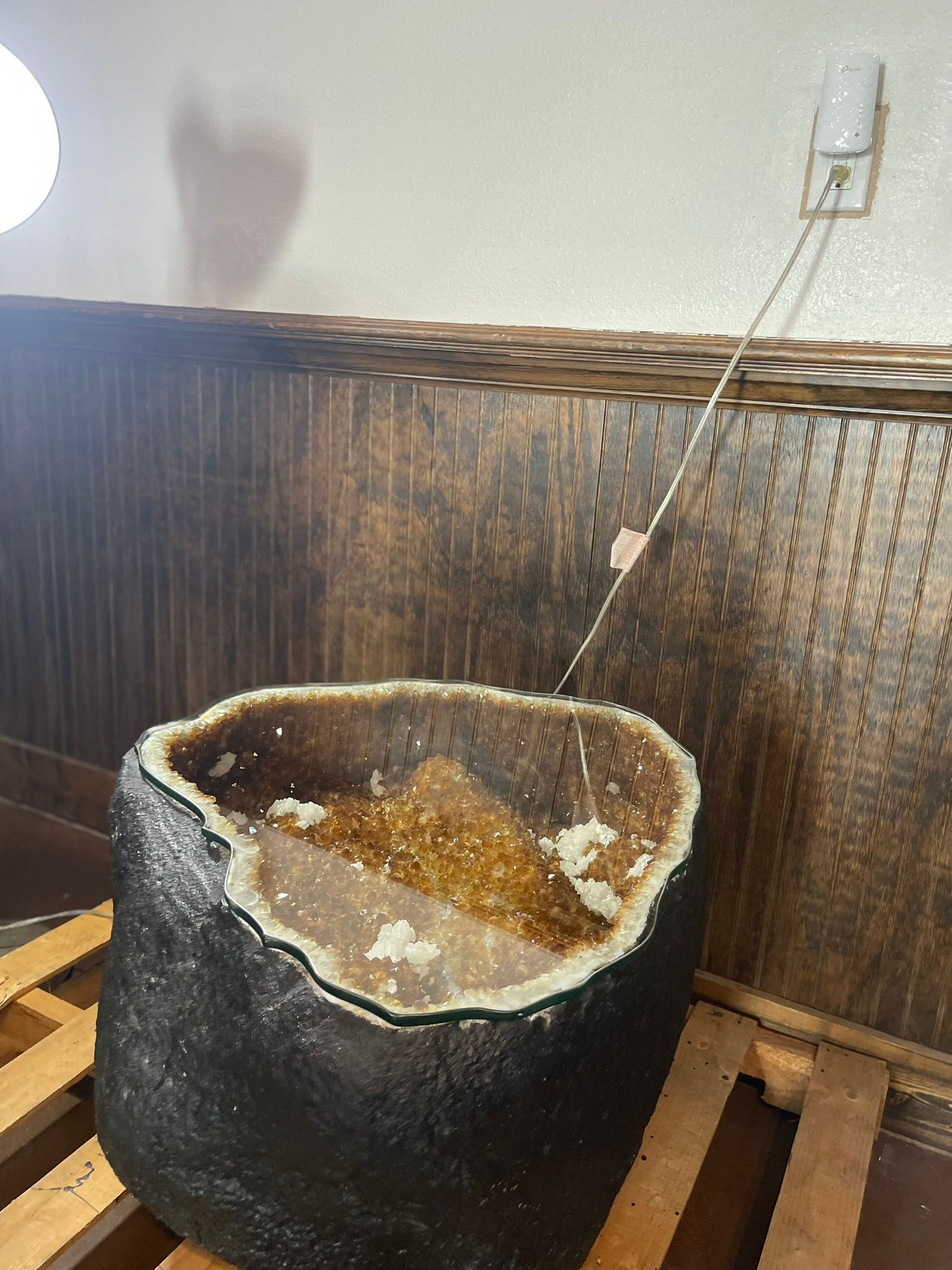 A large geode rock sits on a wooden pallet near a wall with a wall-mounted light shining directly into its crystal center.