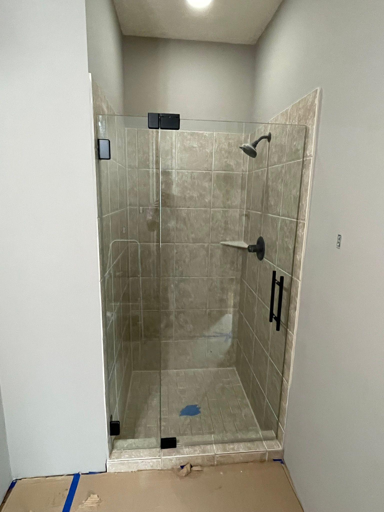 A walk-in shower with a glass door, matte black fixtures, and light-colored tile walls in a partially finished bathroom.