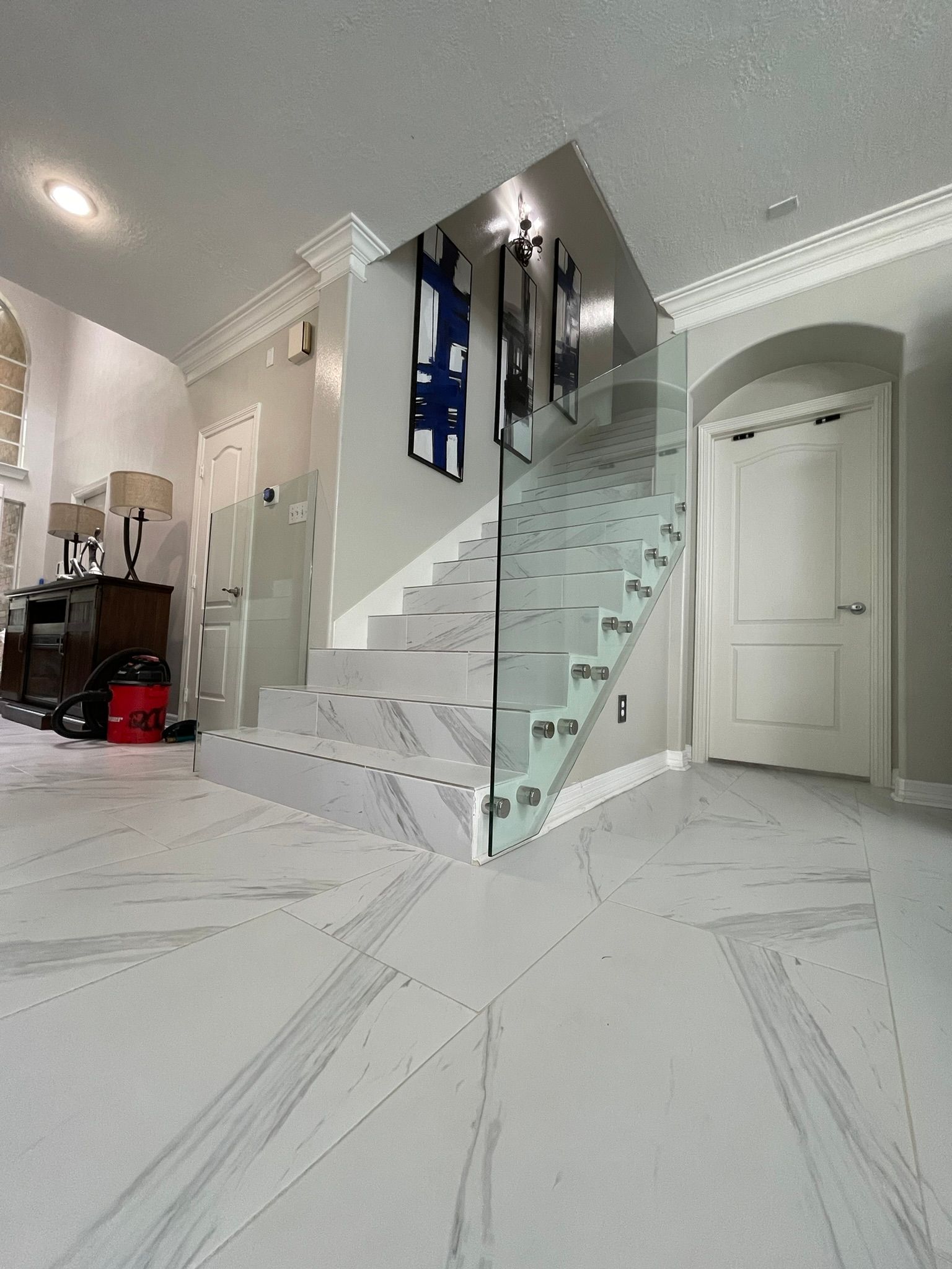 A staircase with white tiled steps, a modern glass railing, and dark wall art, set in a white, marble-floored entryway.