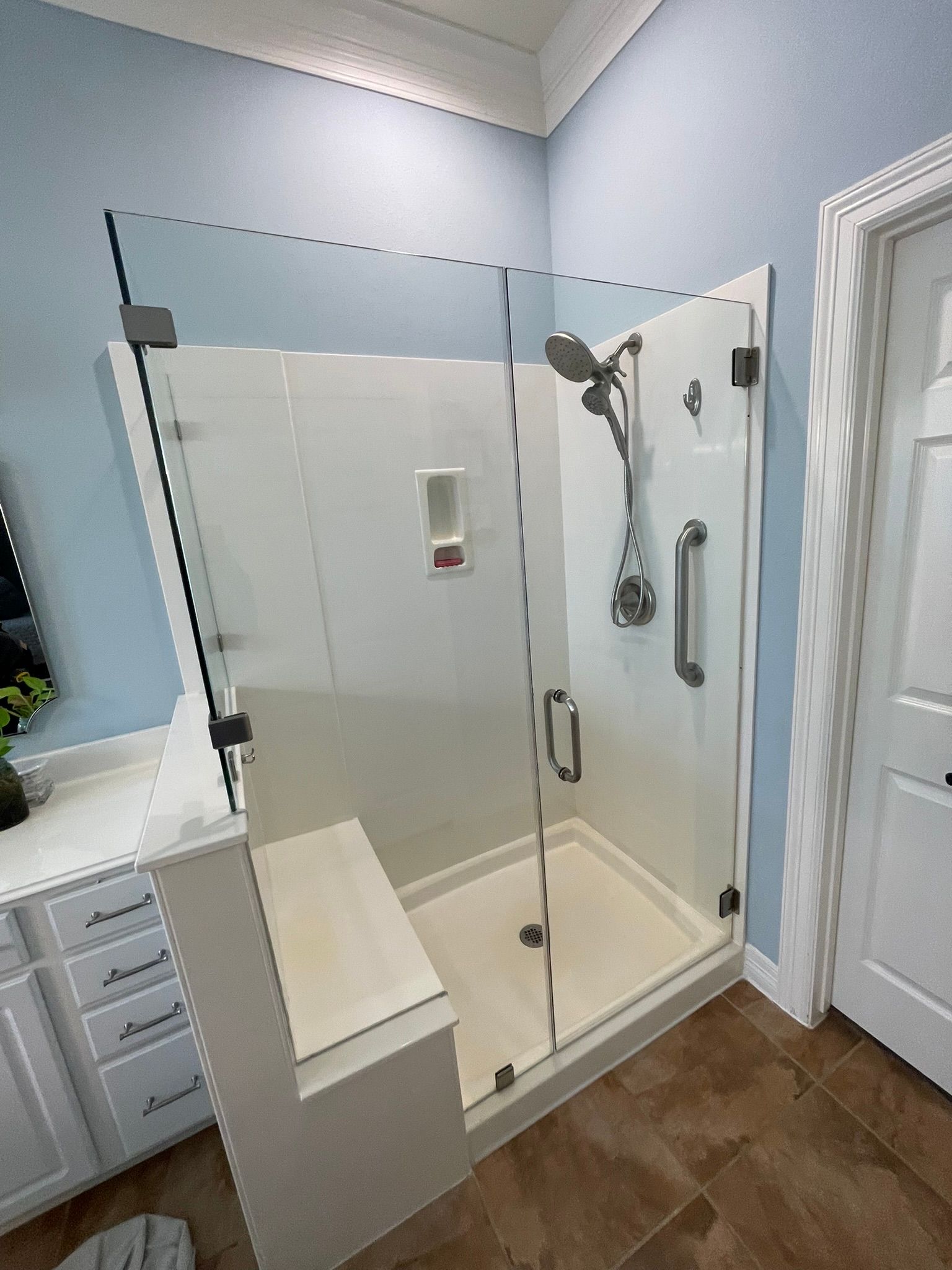 A glass-enclosed shower with a built-in seat, white surround panels, a metal showerhead, and a grab bar.