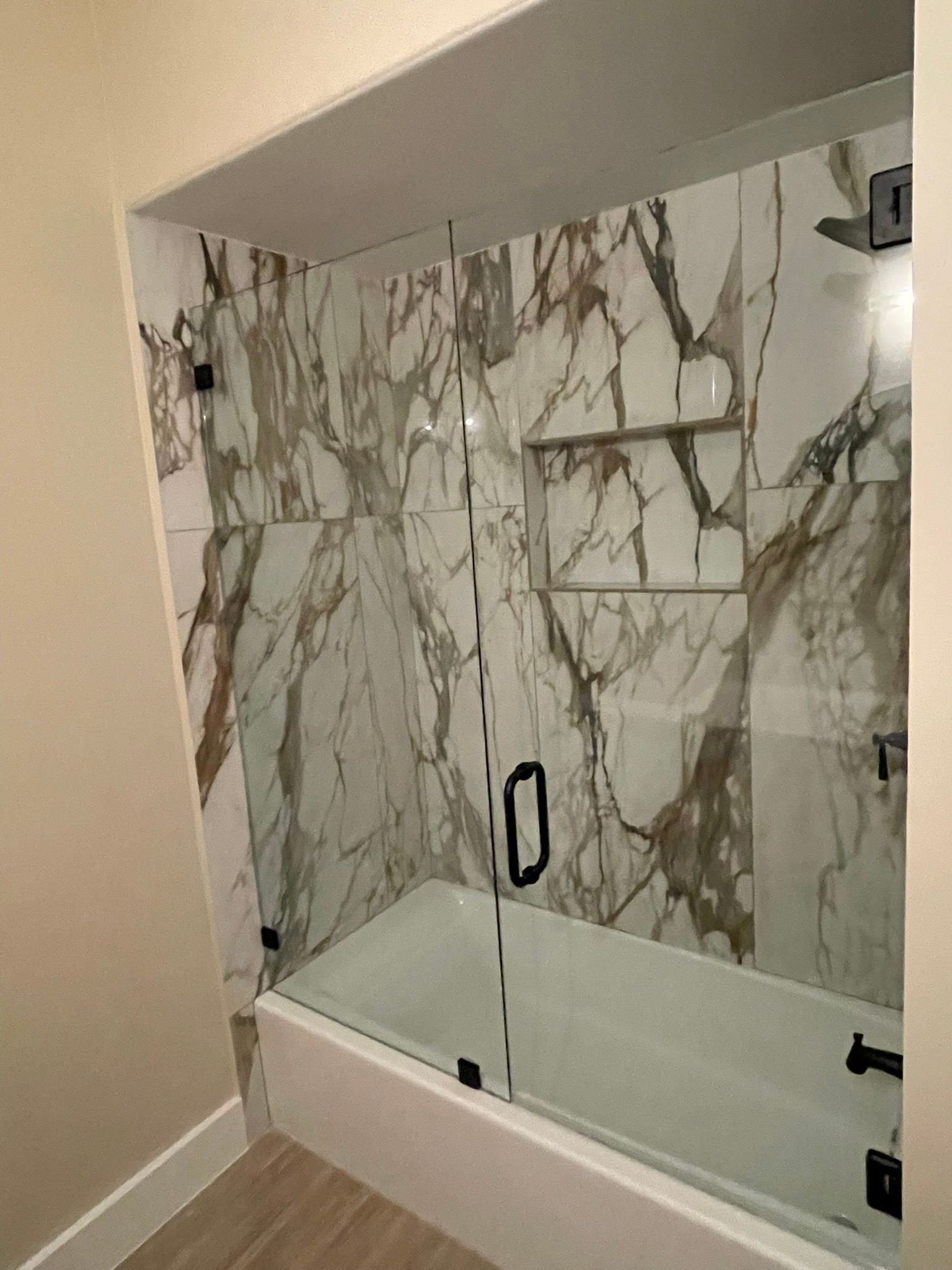 Bathtub and shower alcove with white and gray marble-patterned tile walls, a built-in shelf, and a glass door with black trim.