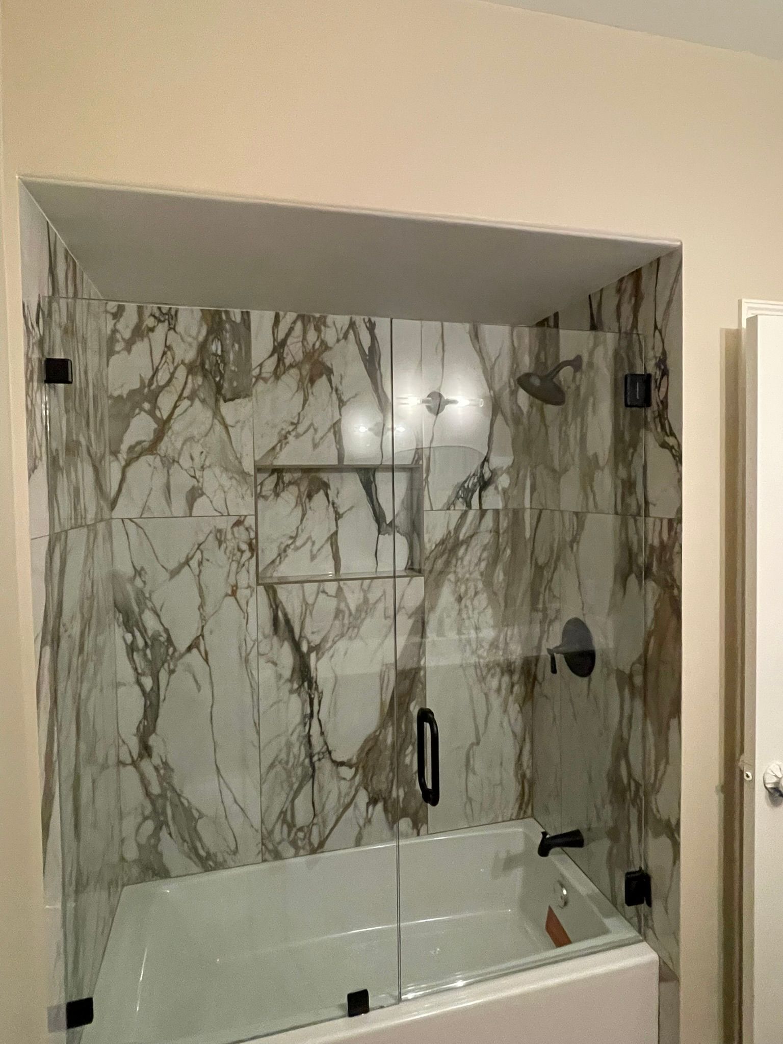 A modern bathtub shower with large-format, brown-veined white marble tiles, glass doors, and matte black hardware.