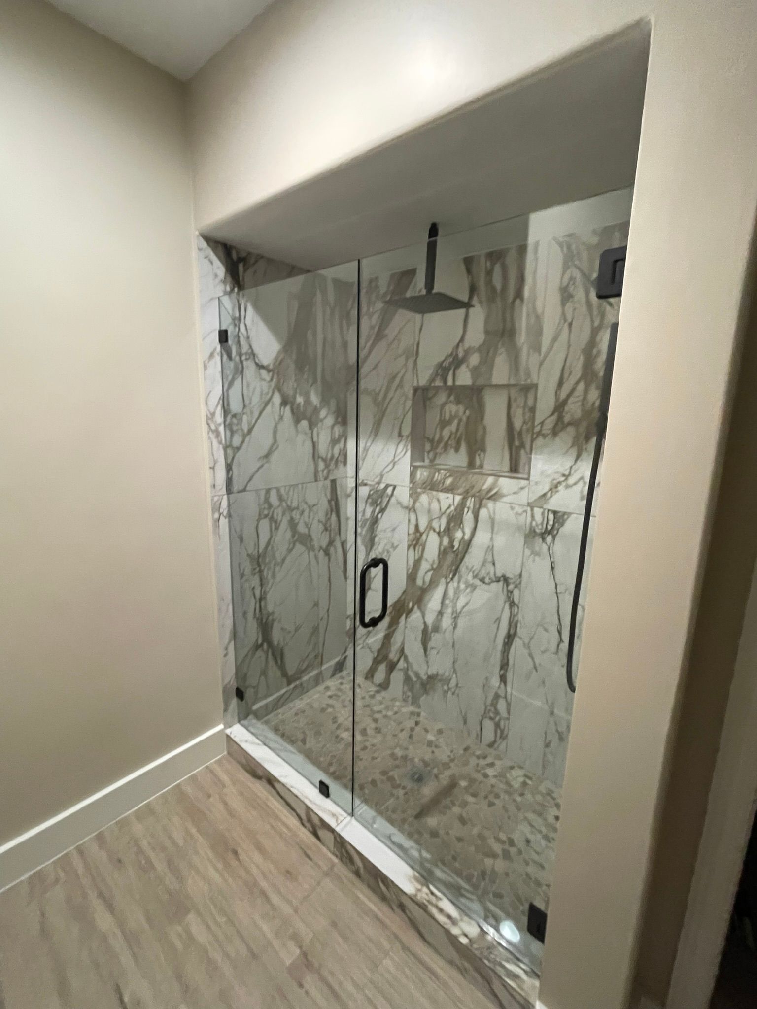 A glass-enclosed shower with marble-patterned tile walls and wood-look flooring in a modern bathroom.