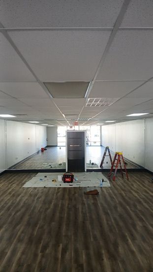 A long, narrow room with wood-look flooring, white walls, and a drop ceiling with two ladders and supplies on a drop cloth.