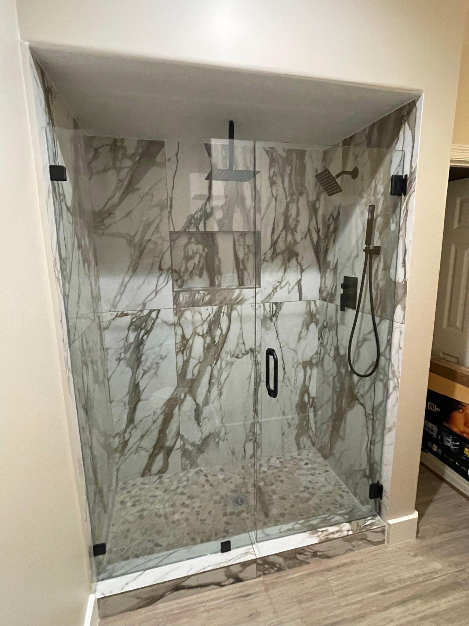 A walk-in shower with white and grey marble tile walls, a pebbled floor, a glass door, and black fixtures.