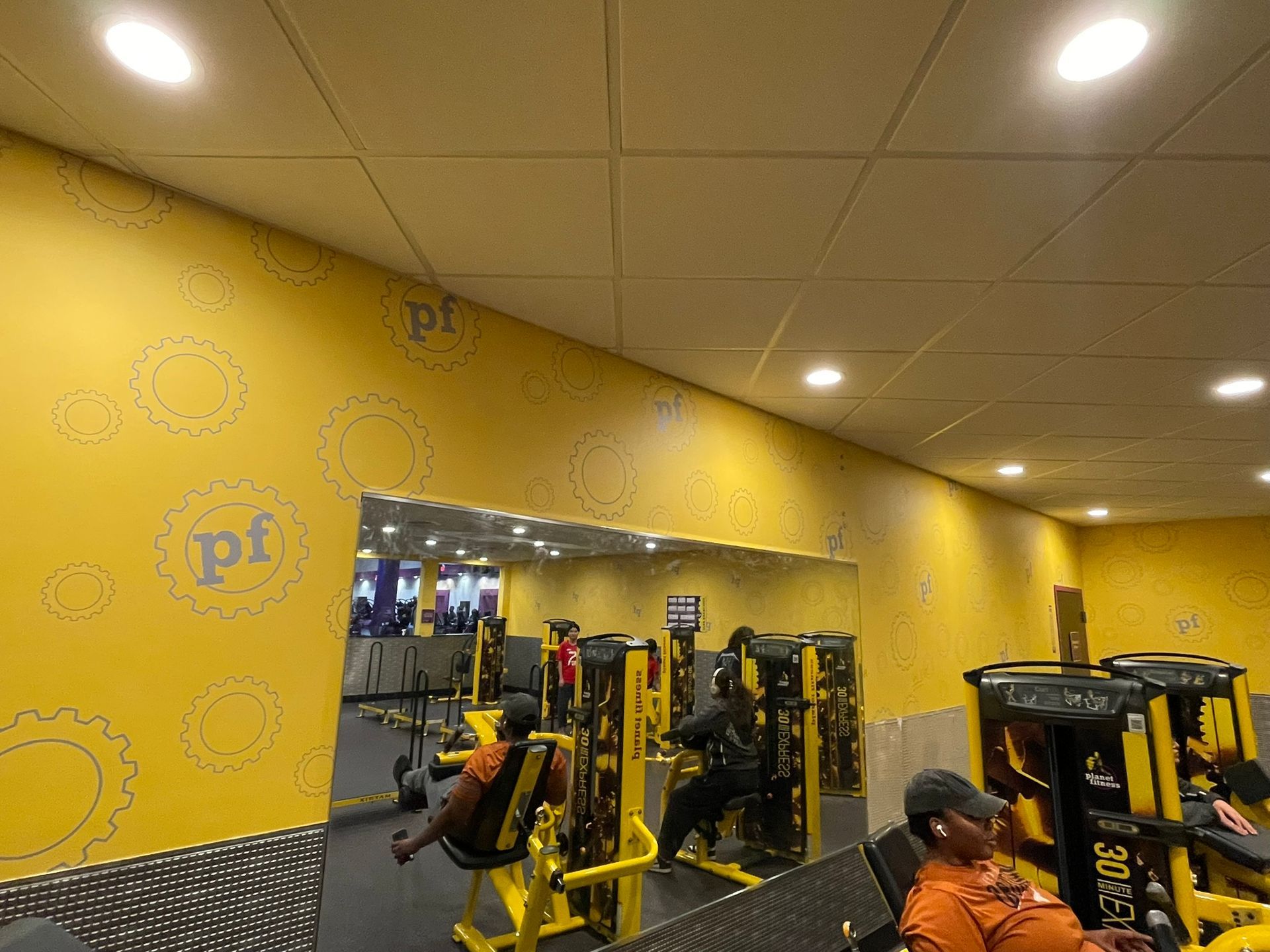 People exercising on yellow gym equipment at a Planet Fitness with yellow walls and large mirrors.