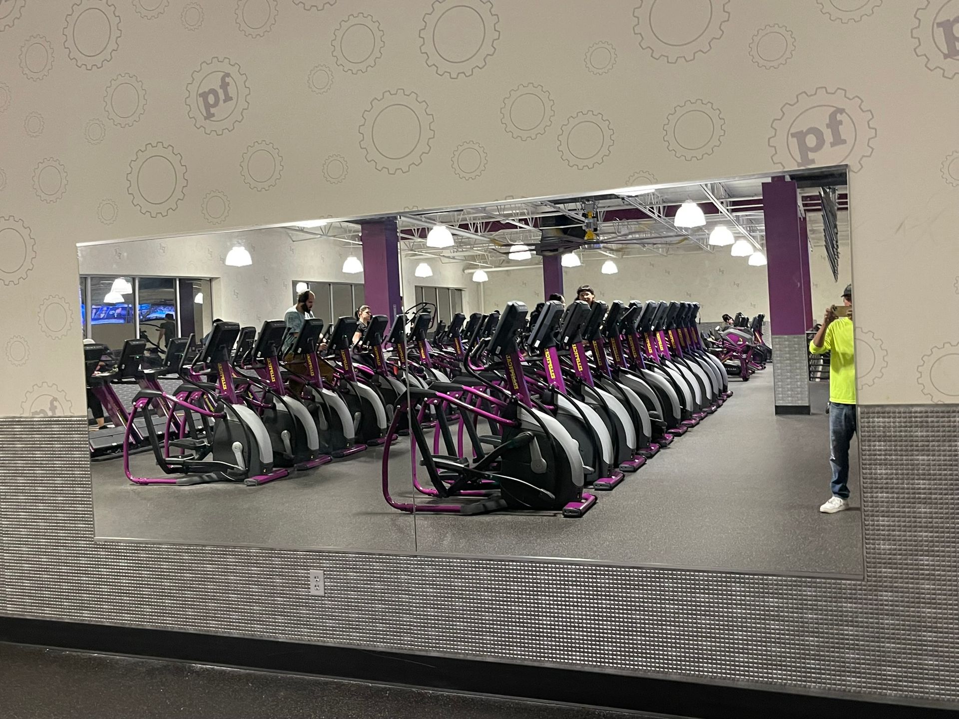 Rows of purple-accented elliptical machines fill a gym, reflected in a large wall mirror with a person standing nearby.