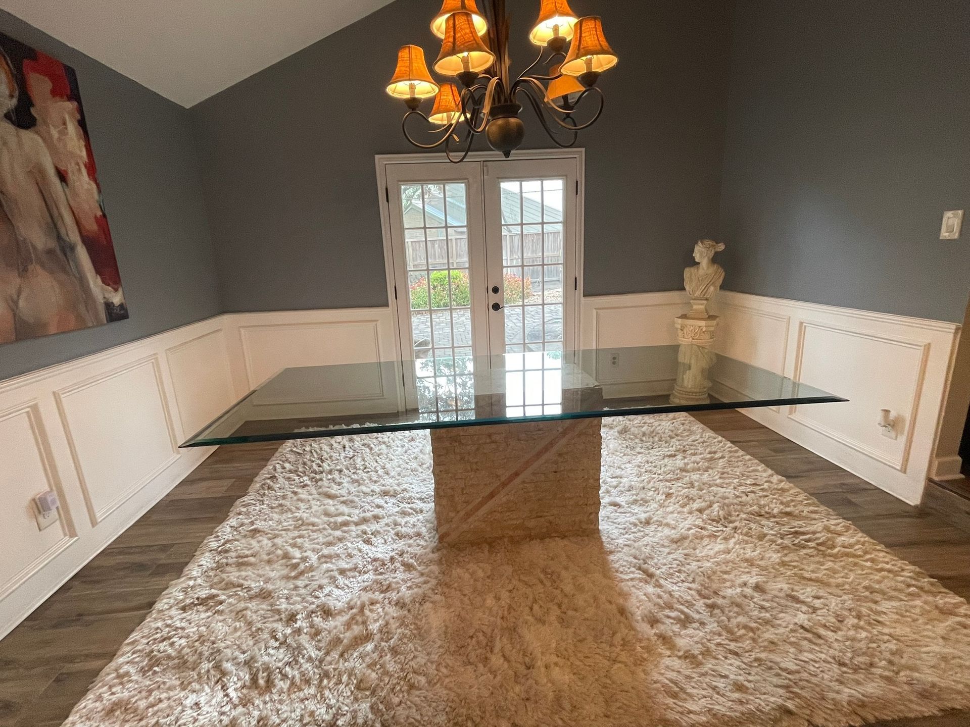 A glass-topped dining table with a stone pedestal base sits on a shaggy rug in a dining room with white wainscoting.
