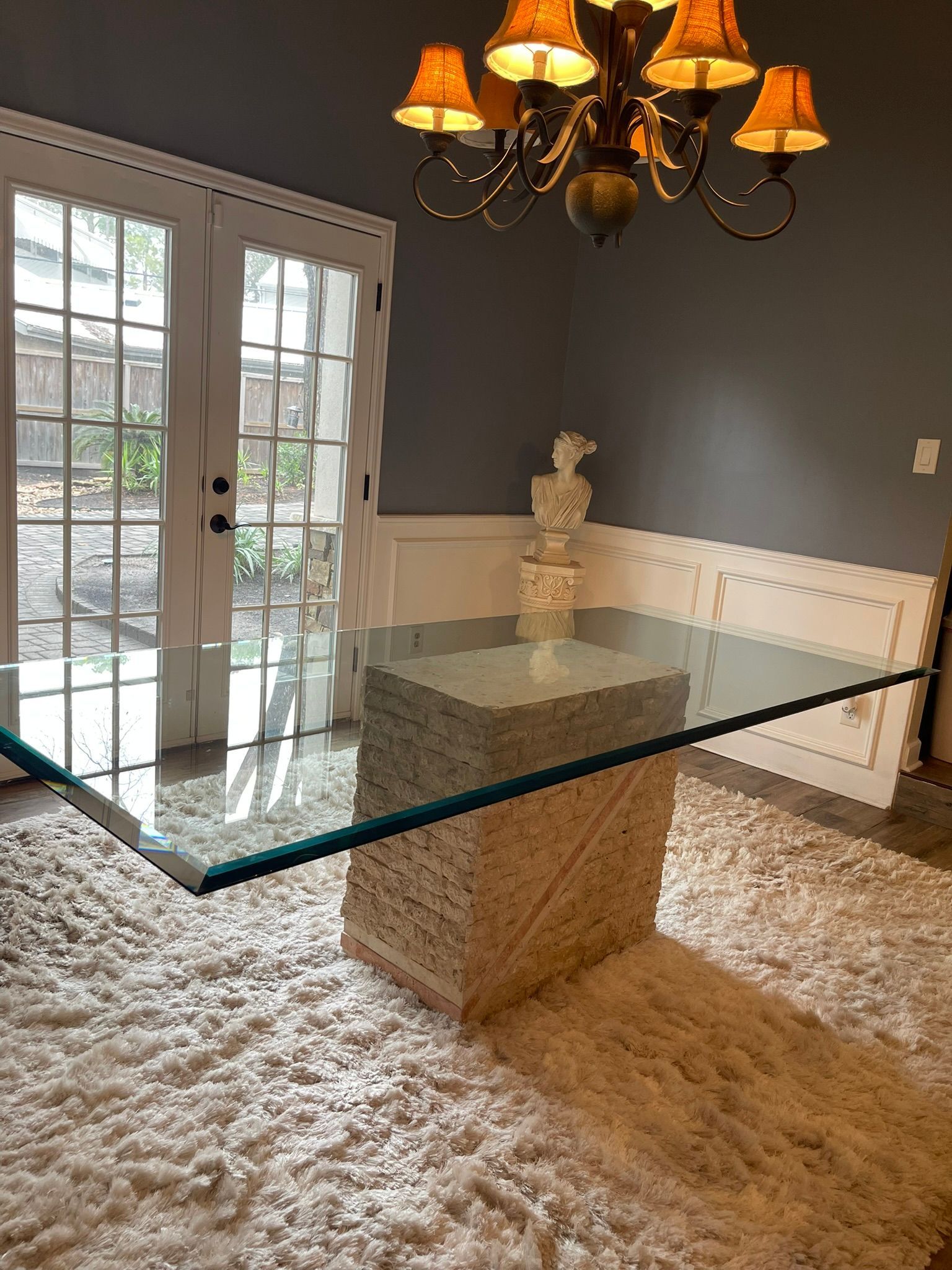 A glass-topped dining table with a stone base sits on a white shaggy rug under a chandelier in a room with gray walls.
