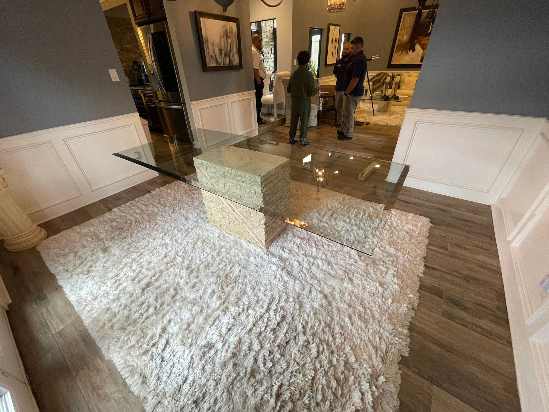 A modern glass table with stacked gold-toned bases sits on a white shaggy rug in a hallway with white paneled walls.