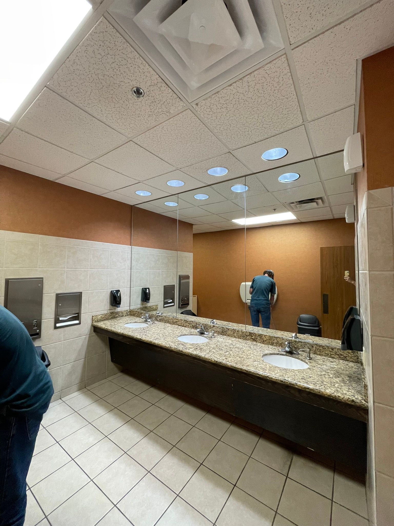 A public restroom with granite countertops, three sinks, and large mirrors on a tan wall above a tiled floor.