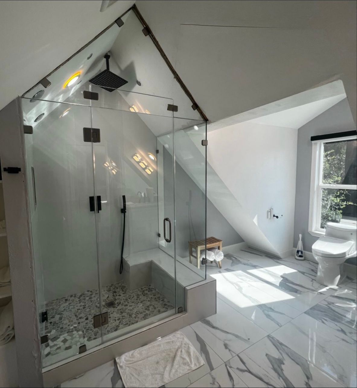 A modern bathroom with a glass-enclosed shower featuring a pebble tile floor, a built-in bench, and white marble flooring.