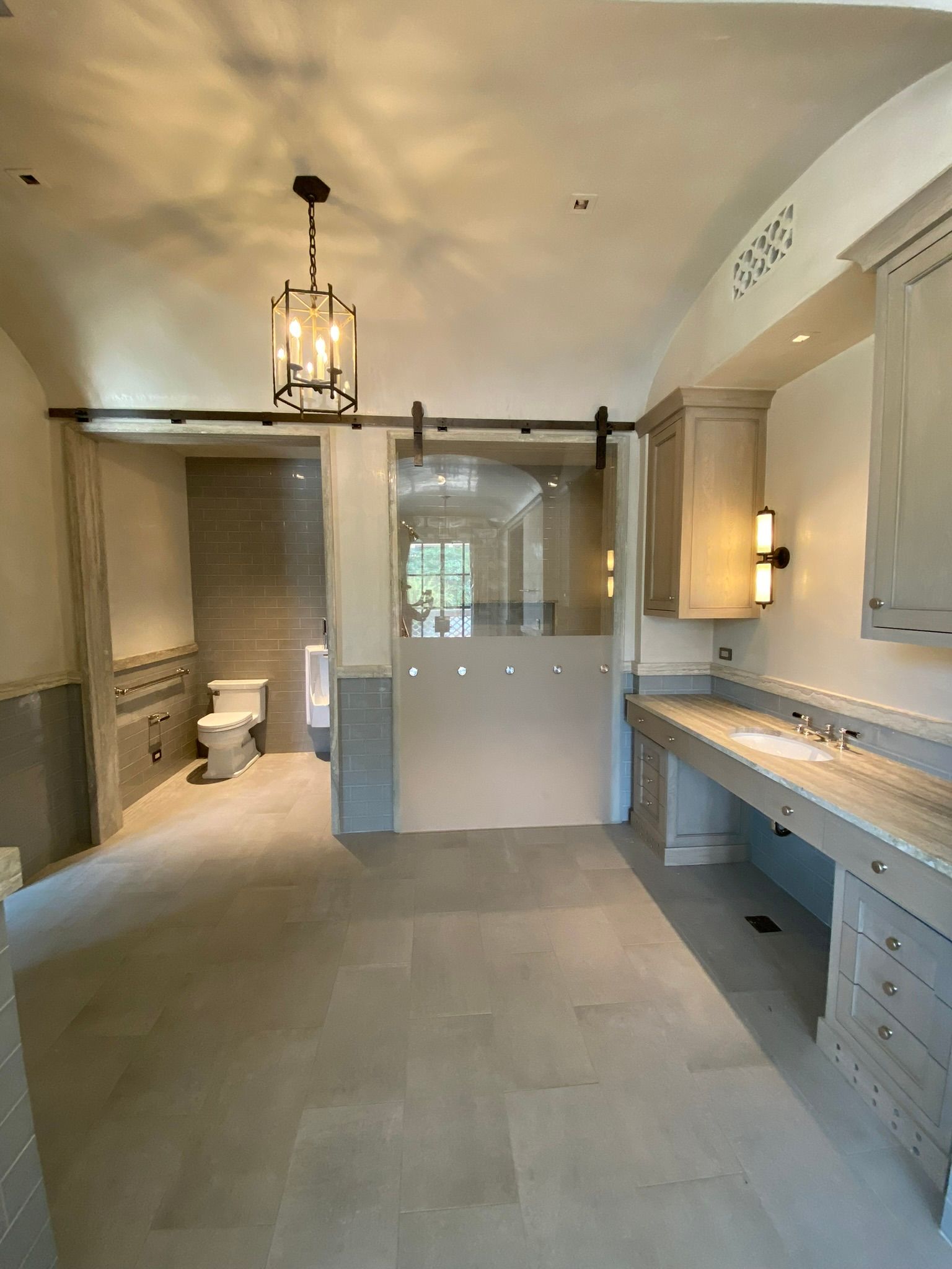 A spacious bathroom with a light-colored tiled floor, a vanity with drawers, a toilet in a niche, and a glass barn door.