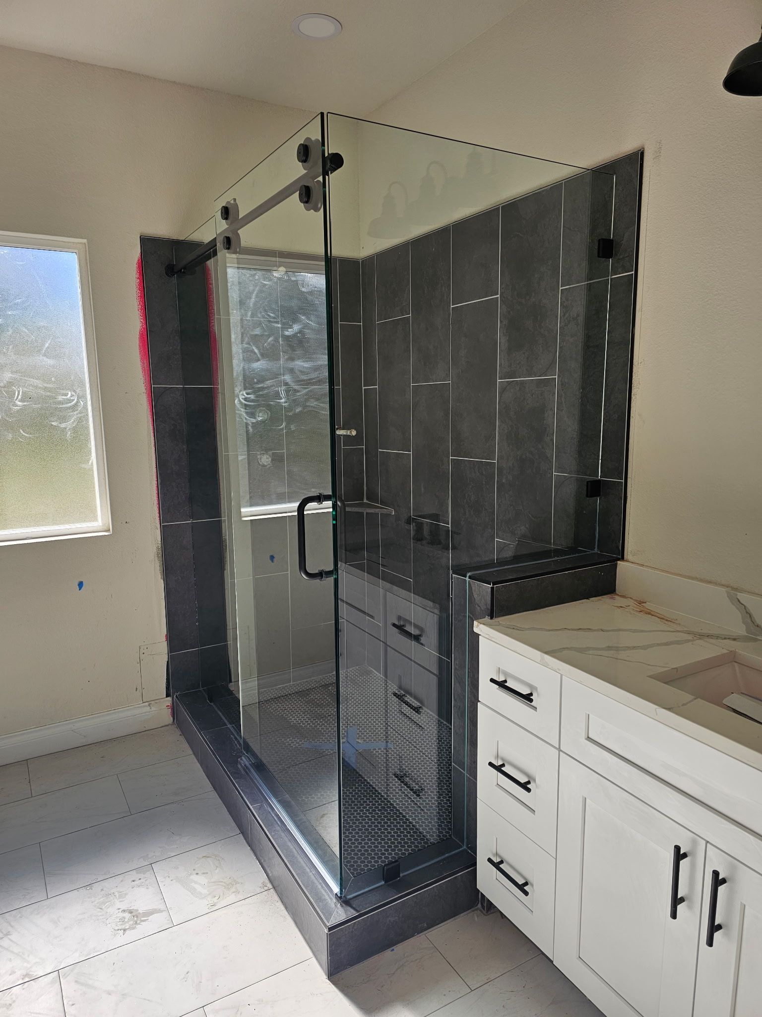 A corner glass shower stall with dark gray tile walls next to a white bathroom vanity with a marble countertop.