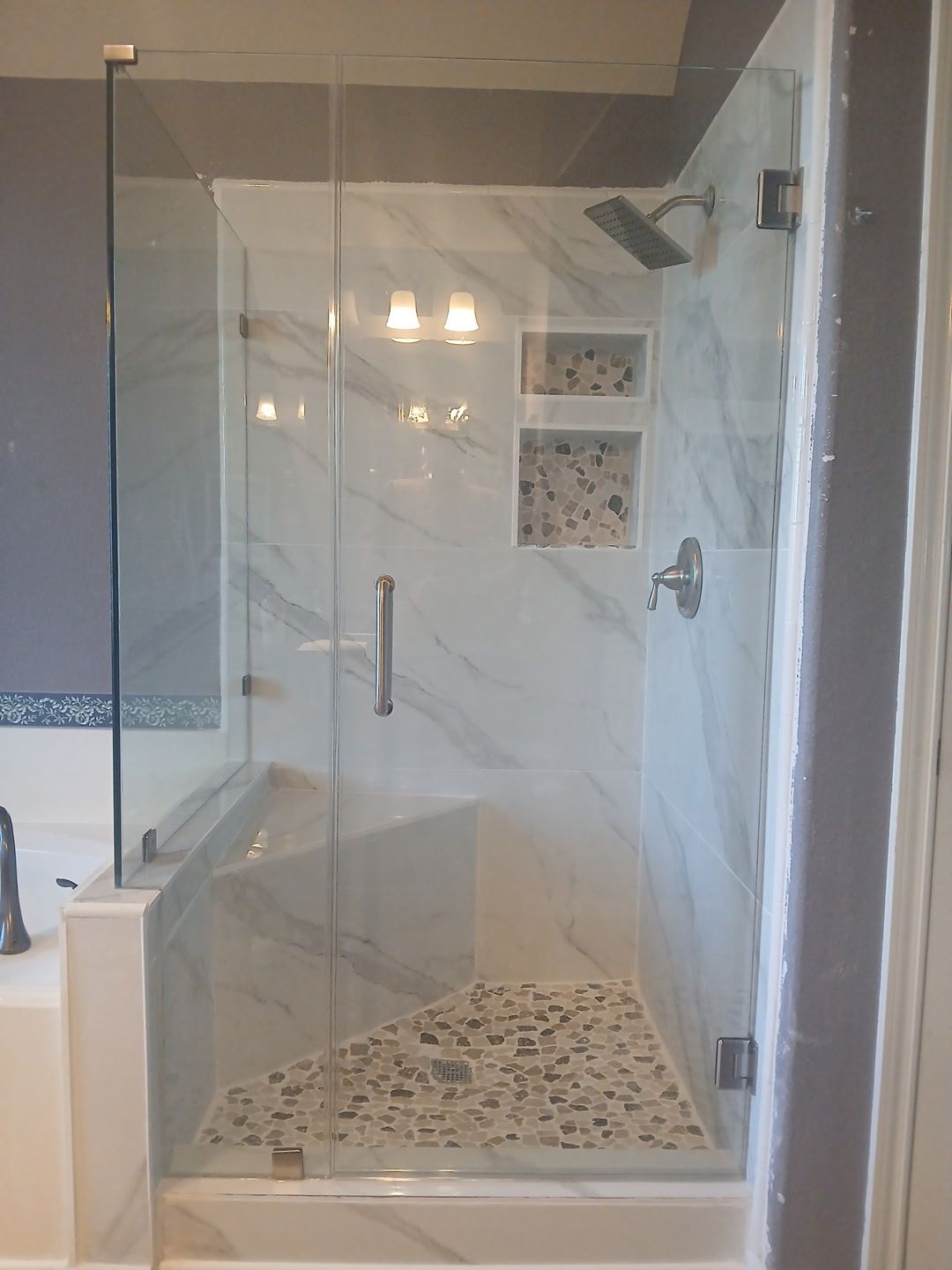 A walk-in shower with white marble-patterned walls, a pebble floor, a built-in bench, glass doors, and dual shower niches.