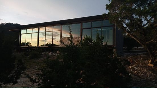 A long, modern house with large glass windows reflecting the sunset sky, surrounded by trees and brush at dusk.
