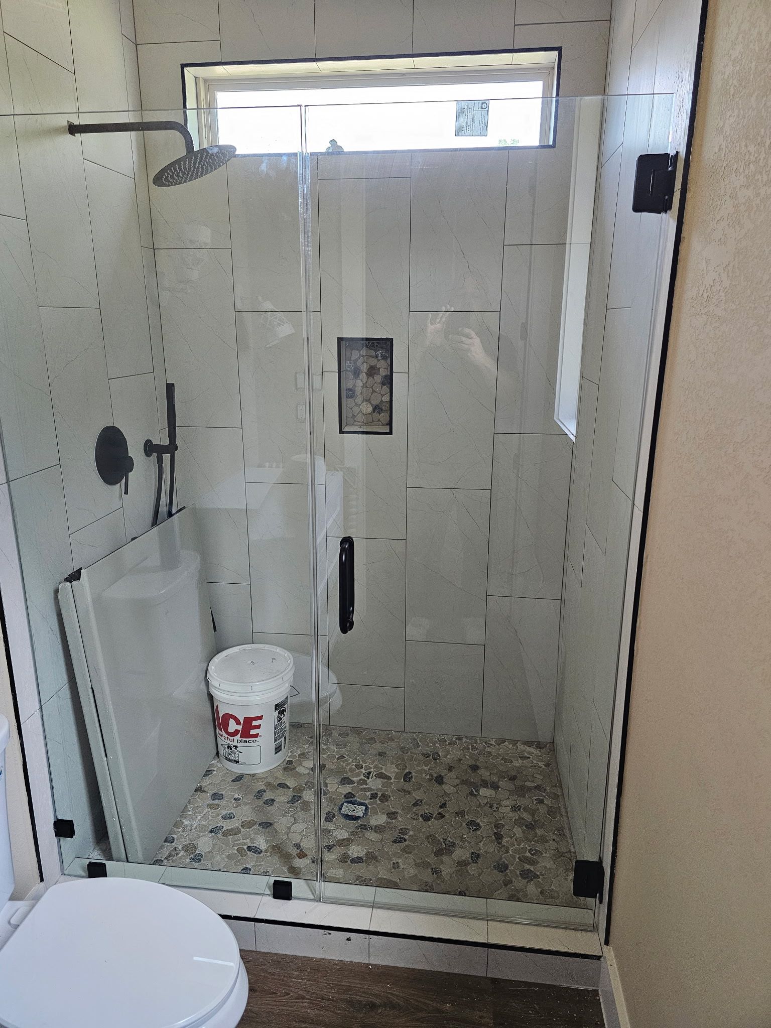 A walk-in shower with a glass door, light-colored tile walls, a pebble floor, a small window, and an ACE bucket inside.