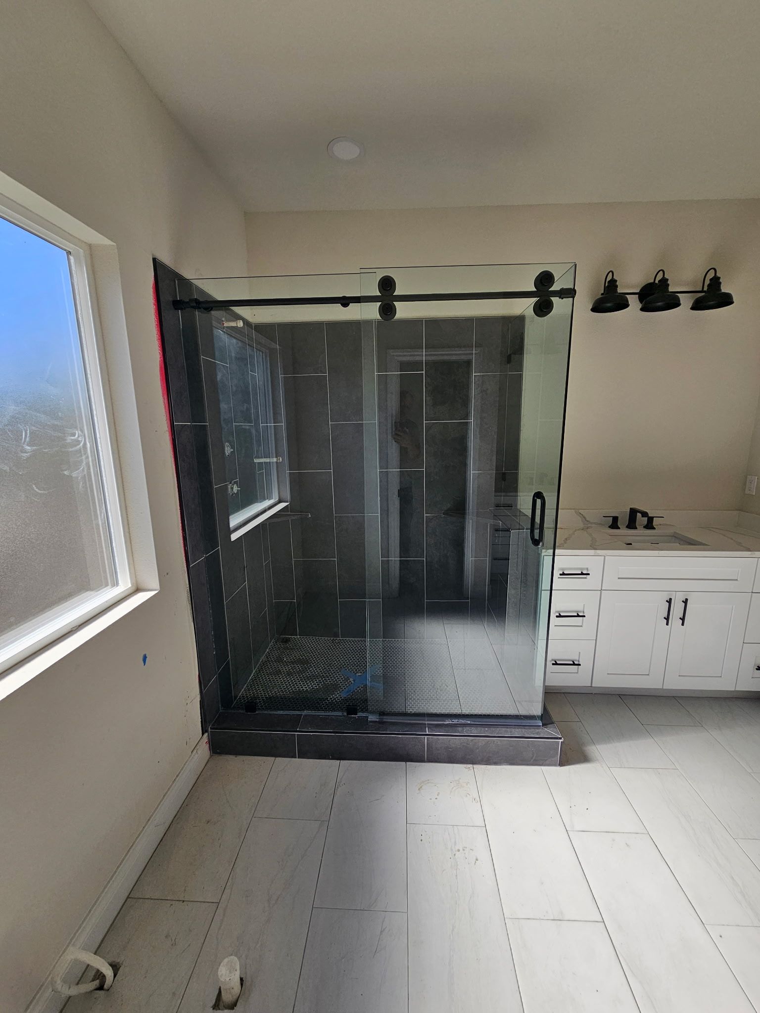A glass shower enclosure with black hardware stands in a bathroom under construction with tiled flooring and a white vanity.