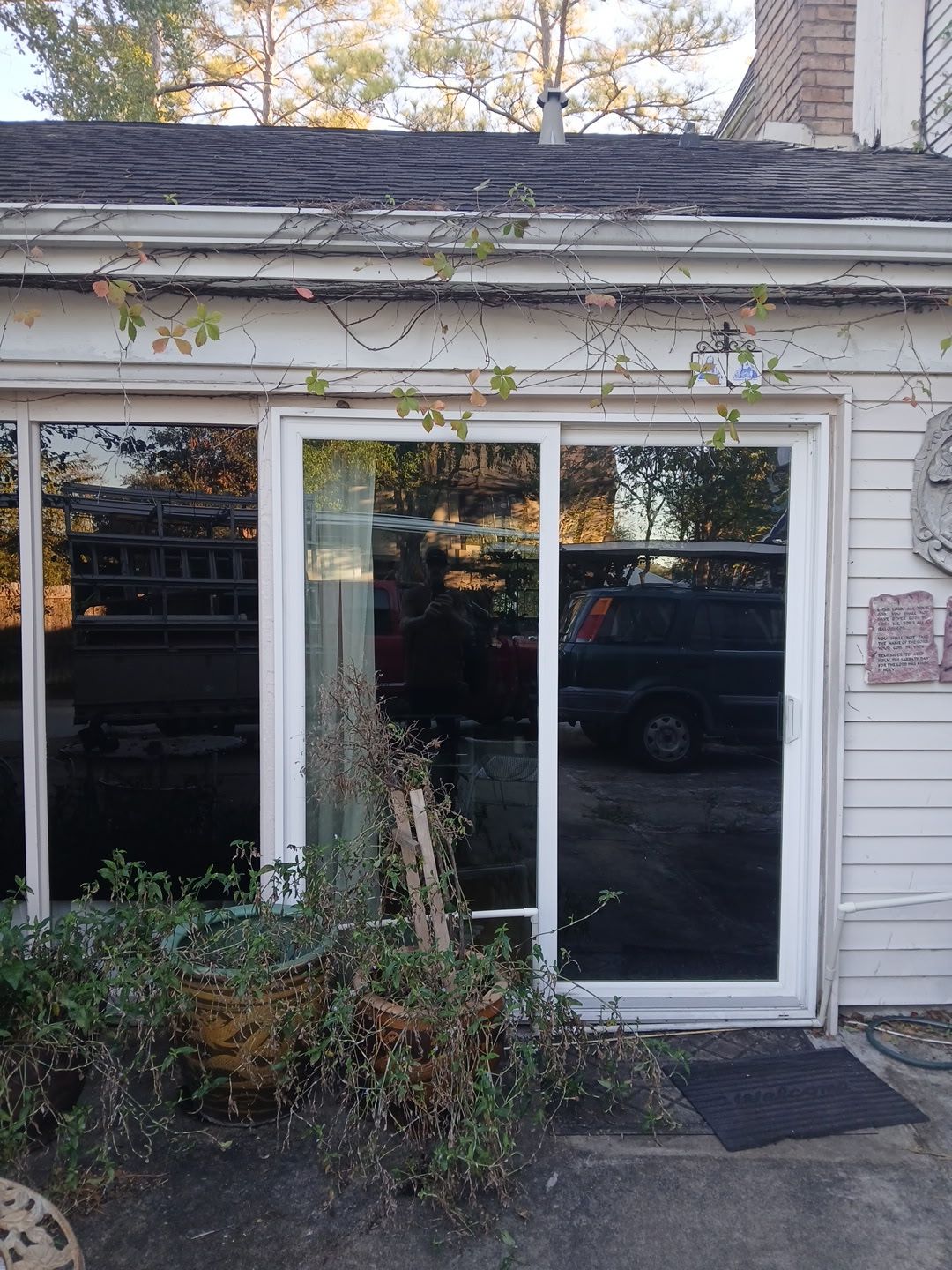 A white sliding glass door on the exterior of a home, framed by horizontal siding and overgrown vines, facing a driveway.