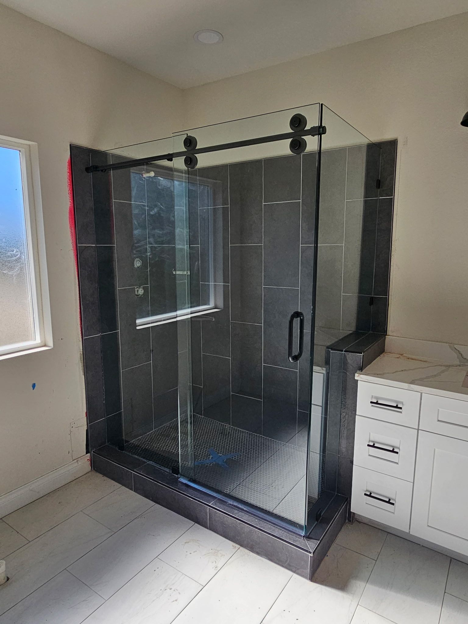 Modern glass shower enclosure with black hardware, installed in a bathroom corner with dark tiled walls and white vanity.