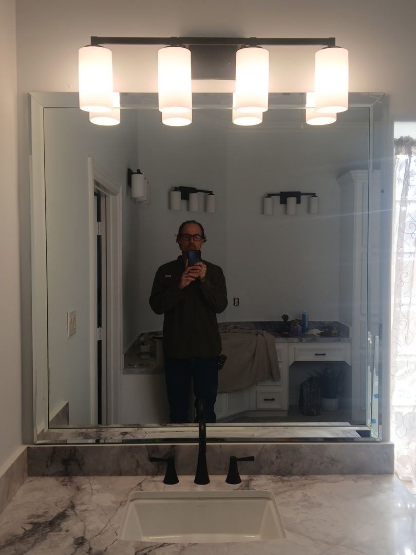 A person standing in front of a bathroom vanity mirror, holding a phone to take a self-portrait.