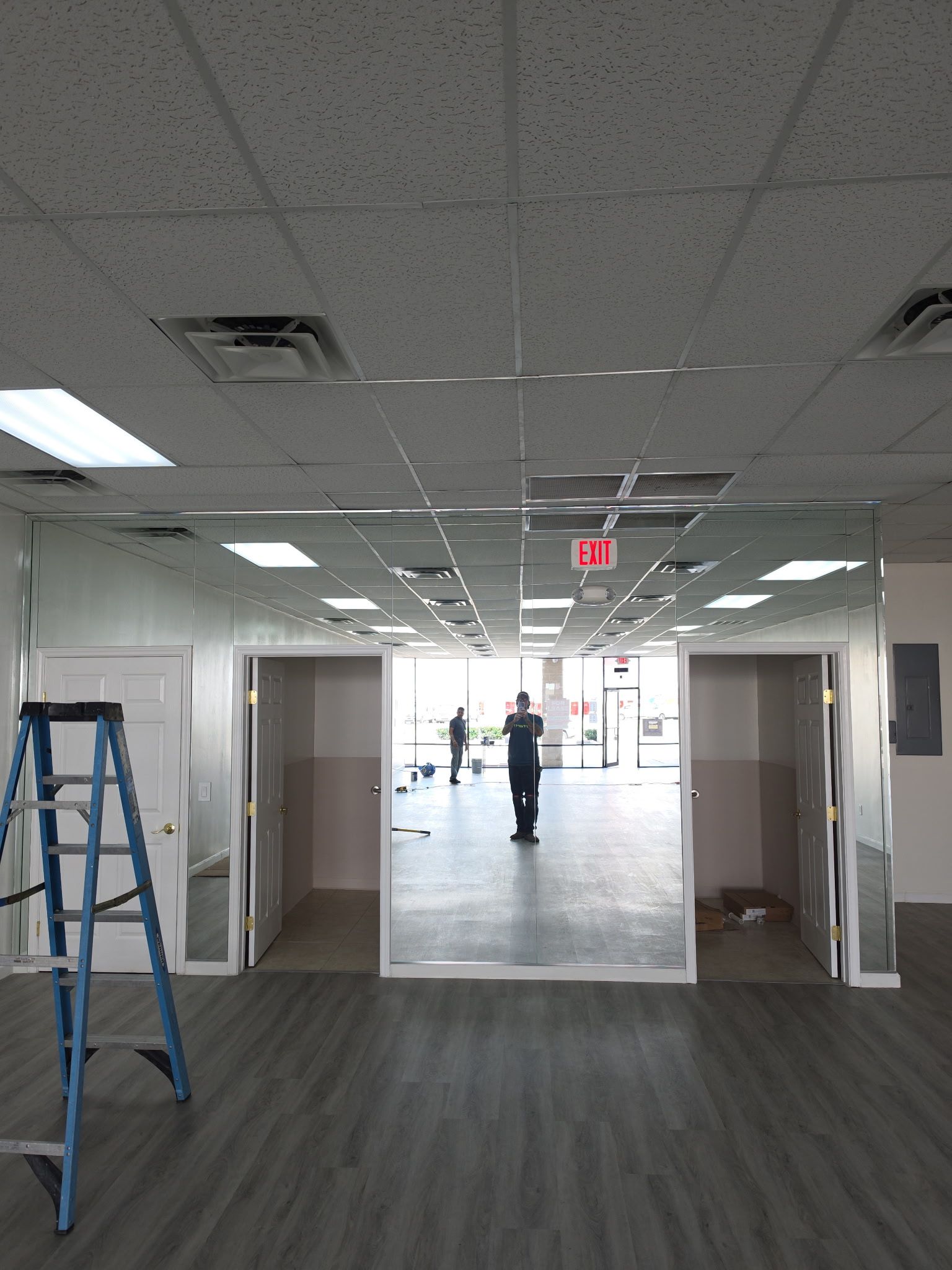 An interior shot of an empty commercial space with a tall ladder, gray flooring, mirrored walls, and open doorways.