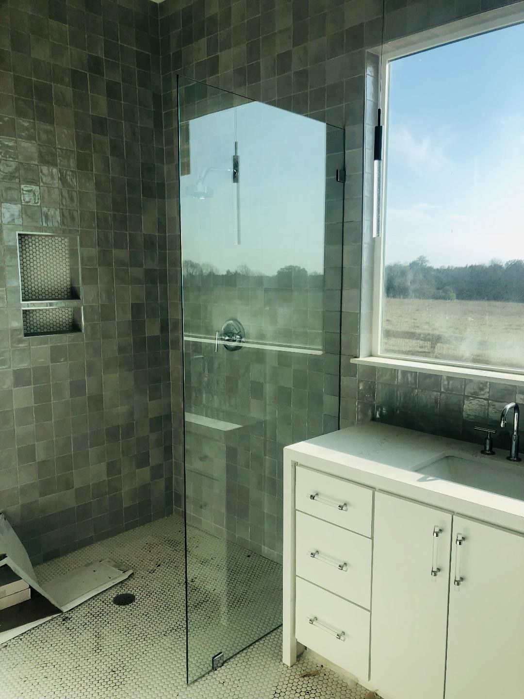 A modern bathroom featuring a glass shower, gray tiled walls, and a white vanity with a countertop sink by a large window.
